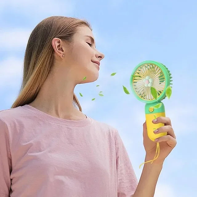 Woman enjoying cool breeze from portable handheld fan with green leaves blowing, perfect for summer outdoor use
